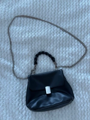 Svart handväska med kedjerem - Snygg svart handväska i slätt konstläder med silverfärgad kedjerem och ett flätat handtag. Väskan har ett lock med rektangulärt spänne i metall och en mjuk, rundad form. Perfekt accessoar för att lyfta din outfit.
