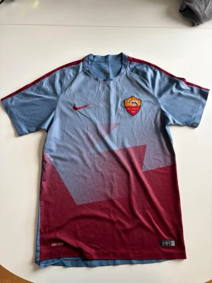 AS Roma blå och vinröd - Snygg AS Roma matchtröja från Nike i blått och vinrött med klubbmärke på bröstet och stora ROMA-trycket på ryggen. Tröjan har korta ärmar, rund hals och är tillverkad i ett lätt, snabbtorkande material med Dri-FIT-teknologi.