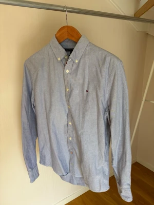 Ljusblå skjorta från Tommy Hilfiger - Klassisk ljusblå skjorta från Tommy Hilfiger med button-down krage och små broderade logodetaljer på bröstet. Skjortan har långa ärmar, knappar framtill och är tillverkad i mjuk bomull. Perfekt för en clean och stilren look.