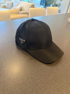Svart keps från Prada i nylon - Snygg svart keps från Prada i glansigt nylonmaterial med klassisk böjd skärm och ikonisk metallogo på sidan. Perfekt accessoar för en trendig och stilren look. Utan prada lapp 