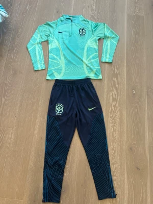 Nike Brazil 🇧🇷 fotbollsträningsset - Säljer ett Nike Brasil fotbollsträningsset med långärmad tröja i ljusblått och gult samt matchande mörkblå byxor med grafiskt mönster. Både tröja och byxor har Brasil-logga och Nike Swoosh. Perfekt för träning eller uppvärmning.
