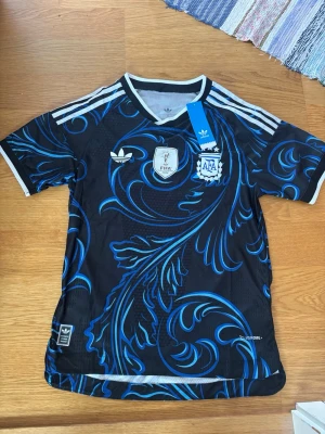 Argentina Adidas fotbollströja svart/blå - Snygg svart fotbollströja från Adidas med blått och vitt mönster, officiella AFA och FIFA World Champions 2022-emblem på bröstet. Tröjan har vita detaljer och klassiska Adidas-ränder på axlarna. Perfekt för dig som älskar fotboll och vill sticka ut på planen.