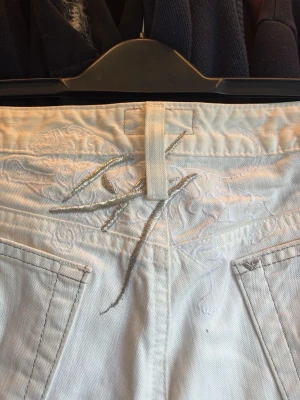 Vita bootcut jeans med silverdetalj - Snygga vita jeans med raka ben men lite bootcut och bakfickor. Diskret broderi baktill. Jeansen har klassisk passform och är tillverkade i jeansmaterial. Perfekta för en clean och fräsch look. Lite småfläckar här och där, mina mammas gamla jeans från Armani. Storlek 27 men skulle säga att det passar mer som 28.