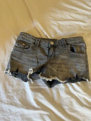 Grå jeansshorts med fransig kant - Säljer ett par grå jeansshorts med rå, fransig kant och slitna detaljer. Shortsen har klassisk femficksdesign, knappgylf och bälteshällor. Perfekta för varma dagar och ger en avslappnad look.