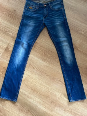 Blå jeans från Superdry - Säljer ett par blå jeans från Superdry med snygg tvätt och raka ben. Jeansen har klassisk femficksdesign, guldfärgade detaljer och Superdry-logga på fickan. Materialet är denim i bomull med lite stretch för skön passform. W32 L34!