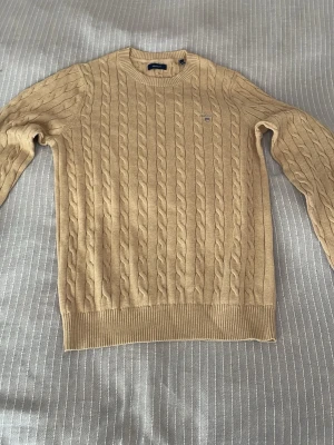 GANT Beige kabelstickad tröja  - Kabelstickad beige tröja från Gant med rund halsringning och ribbade muddar. Tröjan har ett diskret Gant-logomärke på bröstet och är tillverkad i ett mjukt material som passar perfekt för lager på lager. Klassisk och stilren design.
