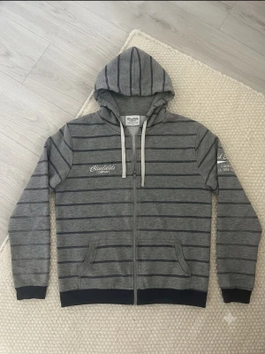 Grå hoodie med blåa ränder Bluefields - Snygg grå hoodie från Bluefields Company med blåa horisontella ränder och broderad logga på bröstet. Dragkedja framtill, två fickor och vit dragsko i huvan. Perfekt för en avslappnad och stilren look.