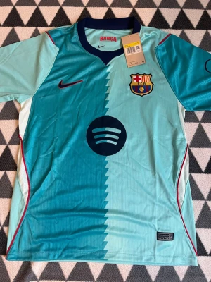 Turkos FC Barcelona matchtröja Nike - Säljer en turkos FC Barcelona matchtröja från Nike med klubbmärke och sponsorlogga på bröstet. Tröjan har korta ärmar, mörkblå krage och ett coolt zigzag-mönster i mitten. Perfekt för dig som älskar fotboll och vill sticka ut på planen.