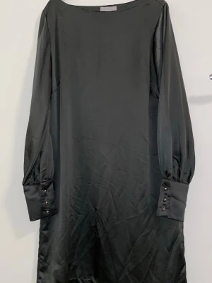 Mörkgrön långärmad satin-klänning - Stilren klänning i satin med lång ärm och breda manschetter dekorerade med knappar. Klänningen har en rak passform och enkel design som ger en elegant look. Perfekt för dig som gillar minimalistisk stil.