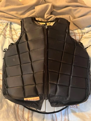 Svart skyddsväst från Racesafe - Säljer en svart skyddsväst från Racesafe med dragkedja framtill och rutmönstrad vaddering. Västen har justerbara sidor och är tillverkad i slitstarkt material, perfekt för ridning eller andra aktiviteter där extra skydd behövs.