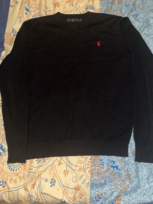 Svart tröja från Ralph Lauren - Klassisk svart tröja från Ralph Lauren med V-ringning och röd broderad logga på bröstet. Tröjan har långa ärmar och ribbade muddar vid ärmslut och nederkant. Perfekt för en stilren look.