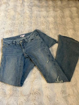 Ljusblå bootcut jeans - Säljer ett par ljusblå bootcut jeans med slitningar på framsidan och snygg broderad detalj på bakfickorna.