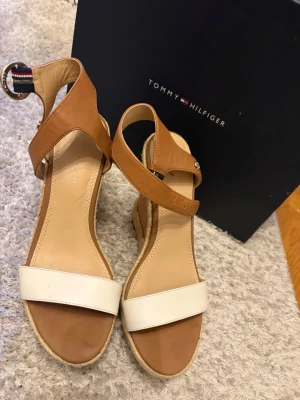 Bruna och vita kilklackssandaler - Snygga sandaler från Tommy Hilfiger med kilklack i rep och läderremmar i brunt och vitt. Justerbart spänne med märkets klassiska banddetalj vid ankeln. Perfekta för sommaren och ger extra höjd med sin stabila platå. Köpta för 1000kr
