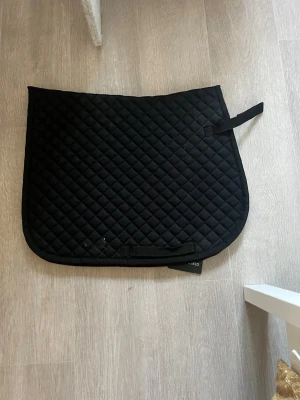 Svart schabrak från FAIRFIELD (OANVÄDT) - Säljer ett svart schabrak med klassiskt rutmönster. Schabraket har kardborreband och är tillverkat i vadderat tyg, perfekt för ridning. Passar till sadel och ger en stilren look till din hästutrustning. 