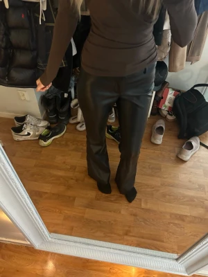 Svarta raka byxor i fuskskinn - Är 170cm lång. Säljer ett par svarta byxor i fuskskinn med utsvängda ben. Byxorna har hög midja och en stilren look som passar till många olika outfits. Materialet ger en cool och trendig känsla. Perfekta för dig som vill sticka ut lite extra. 