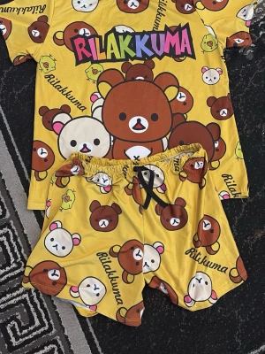 Gul Rilakkuma pyjamas-set - Säljer ett gulligt pyjamas-set med Rilakkuma-tryck i gult, brunt och vitt. Setet består av en t-shirt och shorts med elastisk midja och snörning. Materialet känns mjukt och bekvämt, perfekt för att chilla hemma. Färgglad text och söta björnmotiv över hela plagget.