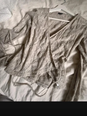 Beige omlottopp i spets - Säljer en beige omlottopp med långa ärmar och vacker spetsdetalj. Toppen har knytband i midjan och en feminin v-ringning. Perfekt för dig som gillar romantiska och trendiga plagg.