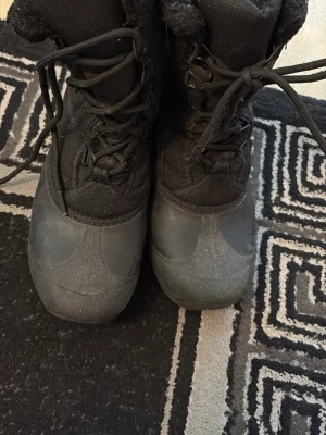 Svarta moon boots med fuskpäls - Säljer ett par svarta moon boots med snörning och fuskpäls på insidan. Skorna har en robust gummisula och ovandel i textil och syntetmaterial, perfekta för kalla vinterdagar. Klassisk moon boot-look som håller dig varm och stilsäker.
