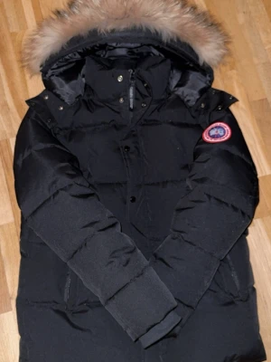 Canada goose  - Snygg svart dunjacka från Canada Goose med klassisk patch på ärmen. Jackan har en stor huva med fluffig pälskant och dragkedja samt knappar framtill. Fylld med dun för extra värme och har två fickor på sidorna. Perfekt för kalla vinterdagar. Pris kan diskuteras 
