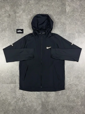 Svart Nike vindjacka med huva - Snygg svart vindjacka från Nike med hel dragkedja framtill och justerbar huva. Jackan har två sidofickor med dragkedja och en vit Nike-logga på bröstet. Tillverkad i lätt och tåligt syntetmaterial, perfekt för blåsiga dagar.