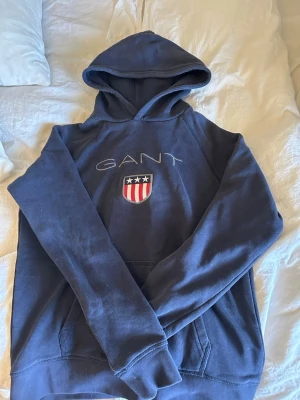 Mörkblå hoodie från GANT med logga - Snygg mörkblå hoodie från GANT med broderad logga och amerikanskinspirerat sköldmärke på bröstet. Klassisk huva och känguruficka framtill. Perfekt för en avslappnad och sportig stil. Tillverkad i mjukt bomullsmaterial.kom privat för att diskutera ett rimligt pris!