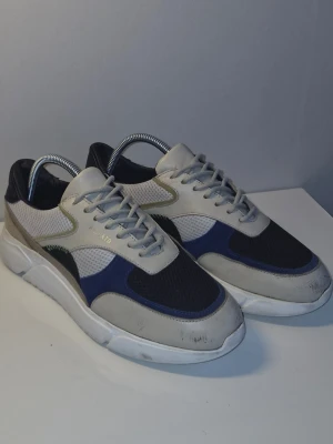 Sneakers från Axel Arigato i blått och vitt - Snygga sneakers från Axel Arigato i vitt, blått och svart med meshpaneler och detaljer i läder. Skorna har en chunky sula och klassisk snörning. Perfekta för dig som gillar streetwear och modern stil.