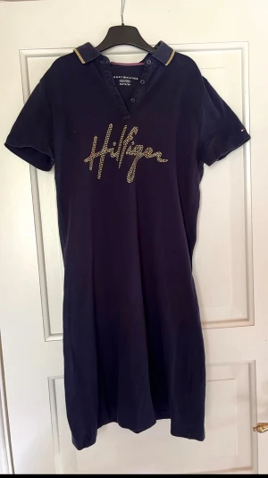 Marinblå pikéklänning Hilfiger - Marinblå pikéklänning från Tommy Hilfiger med korta ärmar, krage med gulddetaljer och Hilfiger-logga i guld på bröstet.