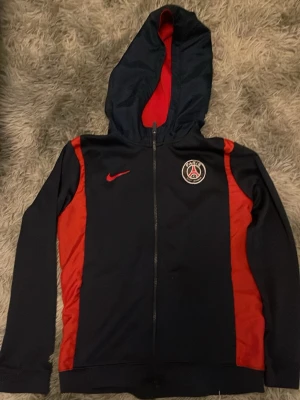PSG hoodie från Nike marinblå/röd  158-170 - Säljer en snygg PSG hoodie från Nike i marinblått med röda detaljer och röd insida på huvan. Dragkedja framtill, PSG-logga på bröstet och Nike Swoosh i rött. Perfekt för dig som gillar fotboll och vill ha en sportig look. Kan sänka pris vid snabb affär  ☺️