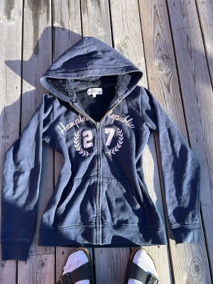 Marinblå hoodie Hampton Republic - Snygg marinblå hoodie med dragkedja från Hampton Republic. Tryck med texten 'Hampton Republic' och siffran 27 framtill, samt vita lagerkransar. Mjuk insida och huva. Perfekt för en avslappnad och sportig stil.