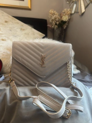 Beige quiltad handväska från YSL - Säljer en beige quiltad handväska från YSL med guldfärgade detaljer och kedjerem. Väskan har ett elegant V-format lock och klassisk YSL-logga framtill. Perfekt storlek för det viktigaste och ger en lyxig touch till din outfit.