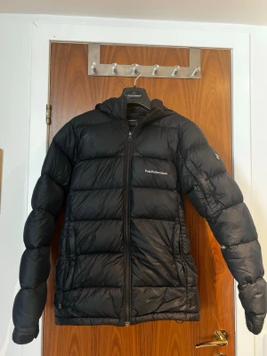 Svart dunjacka från Peak Performance - Säljer en svart dunjacka från Peak Performance med huva och dragkedja framtill. Jackan har två sidofickor med dragkedja och diskret logga på bröstet. Perfekt för kalla vinterdagar tack vare det tjocka dunfodret och den klassiska, puffiga designen. Det finns ett litet hål på vänster arm som är lätt att laga. 