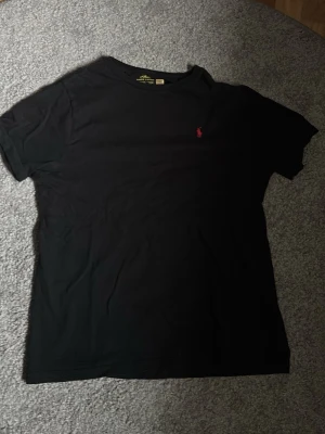 Svart t-shirt från Ralph Lauren - Klassisk svart t-shirt från Ralph Lauren med den ikoniska röda polospelaren broderad på bröstet. T-shirten har rund halsringning och korta ärmar. Perfekt basplagg med stilren design och skön passform. Passar M och S