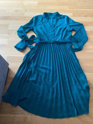 Blå långklänning med plisserad kjol - Snygg blå långklänning med långärmad överdel, ståkrage och knytband i midjan. Kjolen är plisserad för en extra stilren look. Perfekt för dig som vill ha en elegant och trendig klänning.