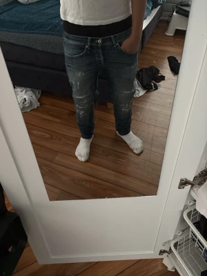 Blå slitna jeans med låg midja - Säljer ett par blå jeans med låg midja och slitna detaljer. Jeansen har smal passform och klassisk femficksdesign. Perfekta för en avslappnad och trendig look. Materialet är denim i bomull.