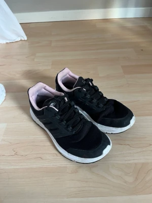 Svarta Adidas sneakers med rosa detaljer - Adidas sneakers med vita sulor och ljusrosa insida. Skorna har klassiska tre ränder på sidan, mesh- och syntetmaterial samt snörning framtill.