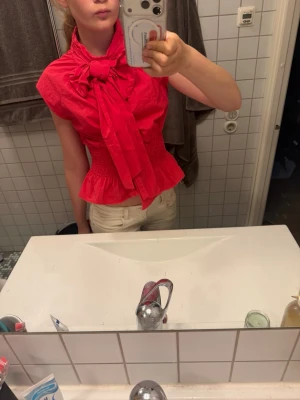 Röd blus med stor rosett i halsen - Snygg röd blus med korta ärmar och en stor rosett i halsen. Blusen har smock i midjan som ger en figurnära passform och volang nedtill. Tillverkad i ett lätt tyg som känns bekvämt och luftigt. Perfekt för dig som vill sticka ut med en färgstark och trendig look. Toppen är lite rosa röd