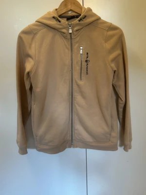 Beige hoodie från Sail Racing - Snygg beige hoodie från Sail Racing med dragkedja framtill och en vertikal bröstficka med zip. Märkets logga är broderad i svart på bröstet. Hoodien har justerbar huva med snören och är tillverkad i mjukt bomullsmaterial. Perfekt för en avslappnad och sportig stil.