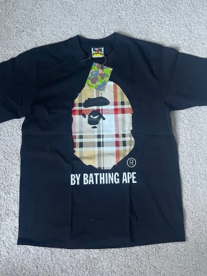 Svart BAPE t-shirt x burburry  - Säljer en svart t-shirt från BAPE med den ikoniska apansiluetten i rutig design på bröstet och texten 'BY BATHING APE' under. Klassisk passform och rund hals. Perfekt för dig som gillar streetwear och vill sticka ut.