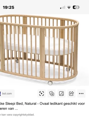 Stokke Sleepi oval spjälsäng i trä - Säljer en Stokke Sleepi spjälsäng i naturligt trä med oval form. Sängen har ribbor runtom, vita hjul för enkel förflyttning och en vit madrass. Perfekt för barnrum och kan anpassas i storlek. Snygg och stilren design.