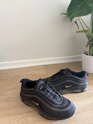 Svarta Nike Air Max 97 sneakers - Säljer ett par svarta Nike Air Max 97 sneakers med guldfärgade swoosh-detaljer. Skorna har den klassiska vågiga designen, synlig Air-sula och är tillverkade i en mix av syntet och textil. Perfekta för dig som gillar streetwear och sportig stil.
