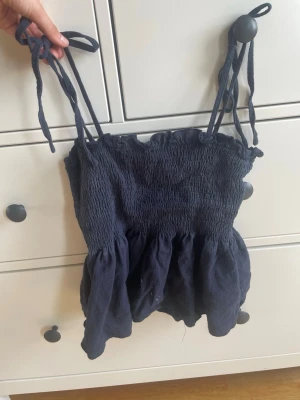 Mörkblå linnetopp med knytband - Supersöt mörkblå linnetopp från LONESS i storlek XS. Toppen har smockad överdel, volangkant upptill och nedtill samt tunna axelband som knyts på axlarna. Perfekt till sommaren och enkel att styla med jeans eller kjol. Pris kan diskuteras 