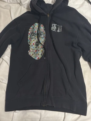 Svart Zip Up Rhinestone - Bvaldi Zip Up Rhinestone Storlek S.   Svart hoodie med dragkedja och huva. På framsidan finns ett stort färgglatt strassmönster av en mask samt texten 'FU' i strass. På ryggen står det 'BANK ROBBER' i stora bokstäver av strass. Hoodien har fickor framtill och en avslappnad passform.