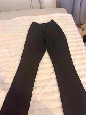 Svarta utsvängda kostymbyxor - Säljer ett par svarta kostymbyxor med utsvängda ben och hög midja. Byxorna har en klassisk look och stängs med knapp och dragkedja framtill. Perfekta för en stilren och trendig outfit.