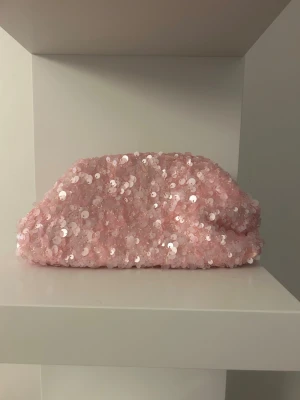 Rosa clutch väska med paljetter - Supersöt rosa kuvertväska täckt av glittriga paljetter. Väskan har en mjuk, puffig form och öppnas upptill med metallspänne. Insidan är fodrad i beige tyg. Perfekt för dig som vill sticka ut med lite extra bling. Och insidan rymmer väldigt mycket!