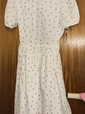 Vit blommig klänning från Kappahl - Supersöt vit klänning från Kappahl med små rosa blommor, puffärmar och öppen rygg. Klänningen har en avslappnad passform, resår i midjan och är gjord i mjuk bomull. Perfekt för sommarens alla tillfällen!