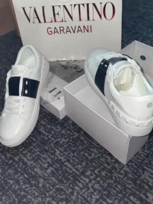 Valentino Garavani vita sneakers - Snygga vita sneakers från Valentino Garavani med bred svart rem över mitten och silverdetaljer. Klassisk låg modell med rund tå och vita skosnören. Tillverkade i slätt läder för en clean och exklusiv look.