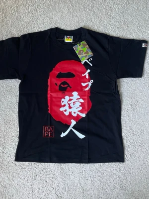 Svart BAPE t-shirt med röd logga - Snygg svart t-shirt från BAPE med stor röd ape-logga och japansk text på framsidan. Klassisk passform och rund hals. Tillverkad i mjuk bomull och har en liten BAPE-logga på ärmen. Perfekt för dig som gillar streetwear och vill sticka ut.