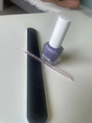Lila nagellack och nagelverktyg - Säljer ett lila nagellack i glasflaska med vitt lock, tillsammans med en svart nagelfil och en glittrig rosa nagelsticka i plast. Perfekt för dig som vill fixa snygga naglar hemma och gillar färgstarka detaljer.