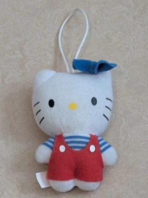 Hello Kitty mjukisdocka med blå rosett - Söt Hello Kitty mjukisdocka med blå rosett på huvudet, randig blå och vit tröja samt röda hängselbyxor. Dockan har en ögla för upphängning och är gjord i mjukt tygmaterial. Perfekt som dekoration eller samlarobjekt.