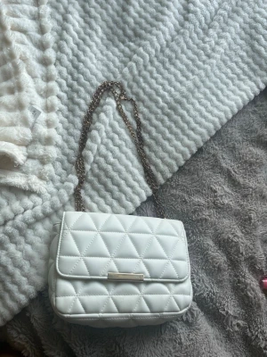 Vit quiltad handväska med kedja - Snygg vit handväska i quiltat mönster med guldfärgad kedjerem. Väskan har en klassisk rektangulär form och ett lock med metallspänne framtill. Perfekt för dig som gillar stilrena accessoarer med en trendig touch.
